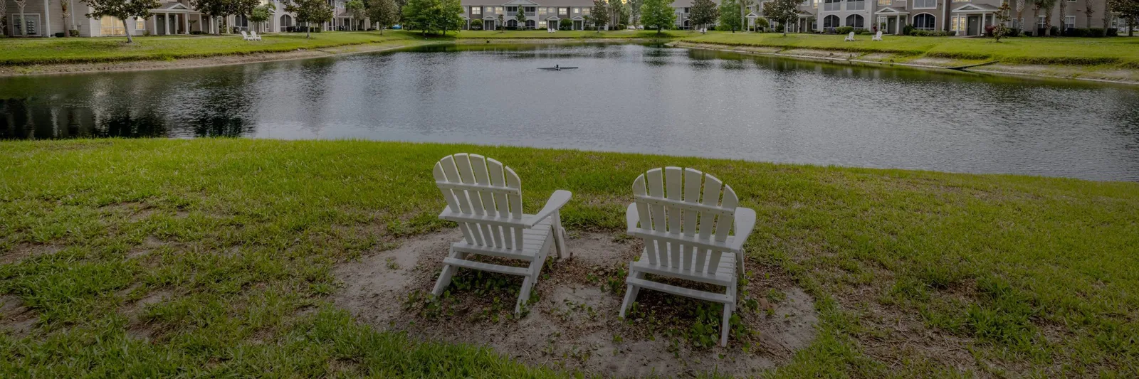 Two white Adirondack chairs on the lawn facing a Pine Lakes pond, with a row of townhomes and a person kayaking in the distance